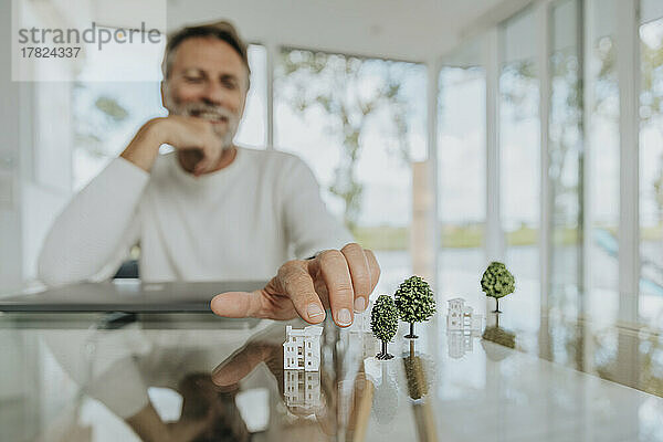 Lächelnder Mann berührt Modellhaus am Tisch sitzend