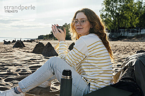 Junge Frau hält Tasse am Strand sitzend