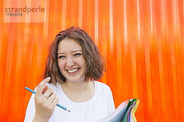 Fröhliches Teenager-Mädchen mit Aktenordnern und Bleistift vor orangefarbenem Frachtcontainer