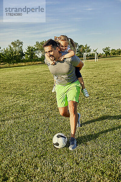 Glücklicher Mann  der Fußball spielt und seiner Tochter Huckepackfahrt auf dem Sportplatz gibt