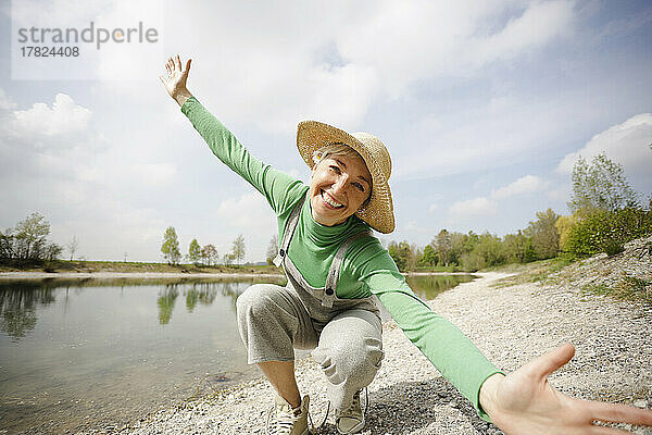 Fröhliche reife Frau mit ausgestreckten Armen genießt den sonnigen Tag am See