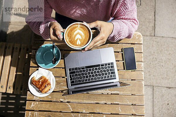 Woman holding coffee cup sitting with laptop at sidewalk cafe on sunny day