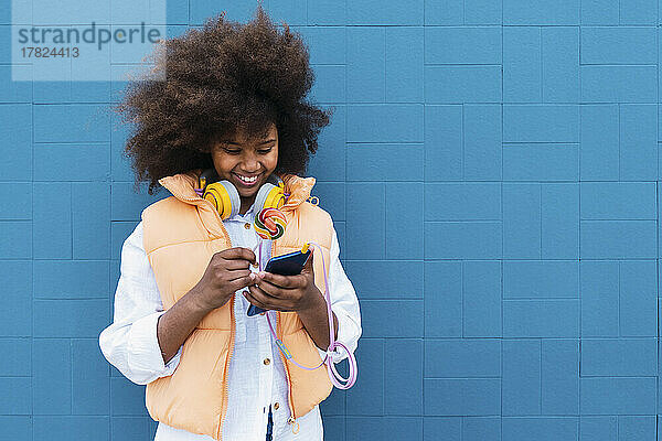 Lächelndes Mädchen hält bunten Lutscher mit Smartphone vor blauer Wand