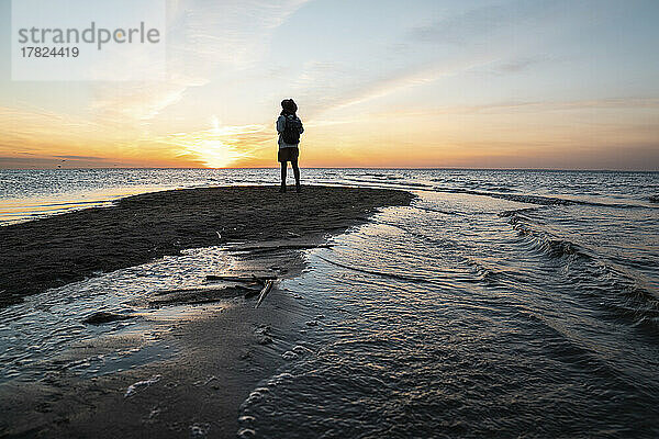 Junge Frau mit Rucksack steht bei Sonnenuntergang an der Küste