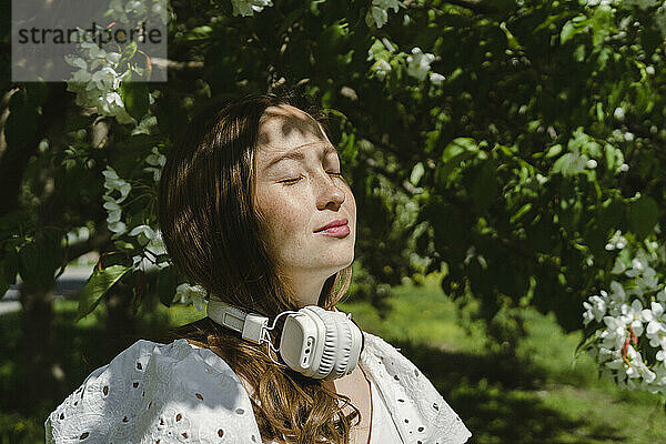 Frau genießt Sonnenlicht mit Kopfhörern um den Hals im Park