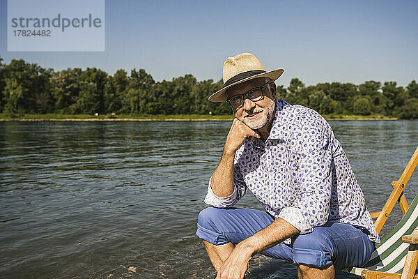 Lächelnder Mann mit Hut sitzt auf Liegestuhl am Flussufer