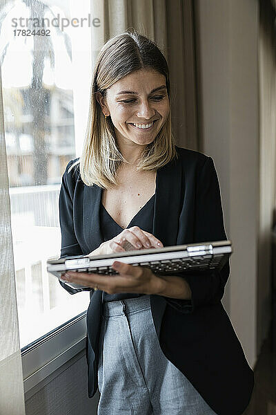 Lächelnde Geschäftsfrau mit Laptop am Fenster des Hotels