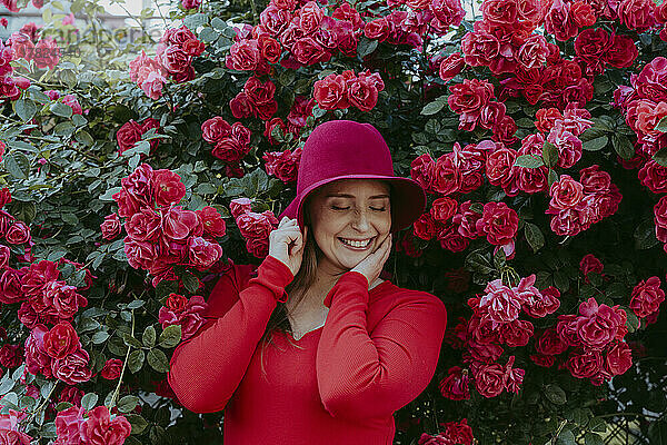 Lächelnde Frau mit rotem Hut steht mit geschlossenen Augen inmitten von Rosen