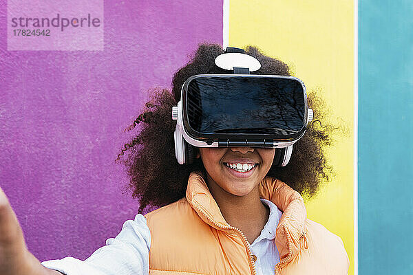 Lächelndes Mädchen mit Virtual-Reality-Simulator  das vor einer bunten Wand steht