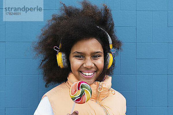 Lächelndes Mädchen mit kabellosen Kopfhörern  das mit Süßigkeiten vor der blauen Wand steht