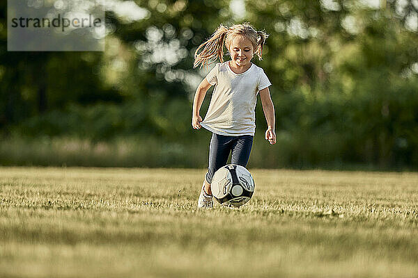 Glückliches blondes Mädchen  das auf dem Sportplatz Fußball spielt