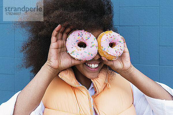 Fröhliches Mädchen bedeckt Augen mit Donuts vor blauer Wand