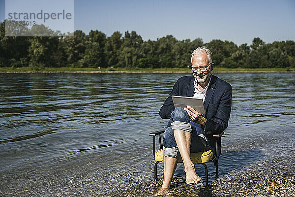 Lächelnder älterer Mann sitzt mit Tablet-PC auf einem Stuhl am Flussufer