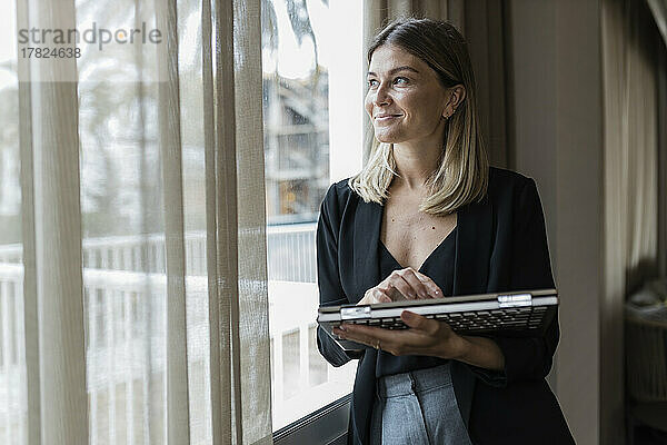 Glückliche Geschäftsfrau mit Laptop und Blick durch das Fenster im Hotel