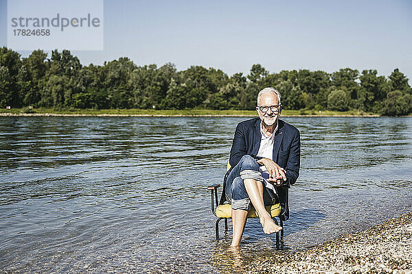 Lächelnder Mann im Blazer sitzt auf einem Stuhl am Flussufer