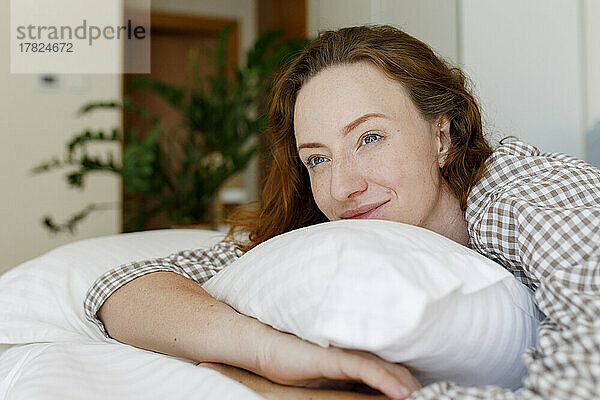 Lächelnde Frau liegt mit Kissen im Schlafzimmer auf dem Bett