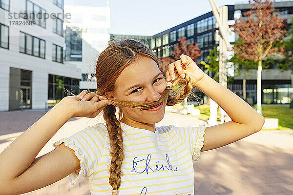 Lächelndes Mädchen  das vor dem Gebäude einen Schnurrbart mit Haaren macht