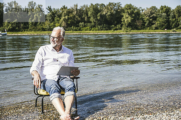 Älterer Mann mit Tablet-PC sitzt auf Stuhl am Flussufer