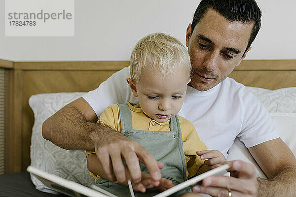 Man reading book with son sitting on bed at home