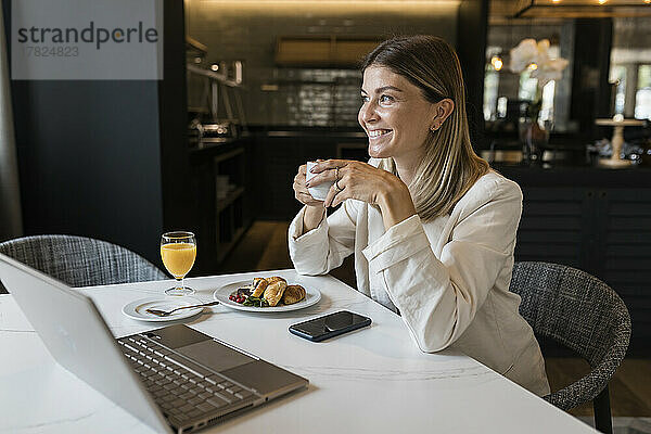 Lächelnde Geschäftsfrau mit Kaffeetasse und Laptop im Restaurant
