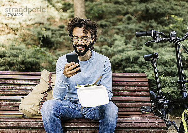 Lächelnder Mann mit Lunchbox und Telefon auf Bank sitzend