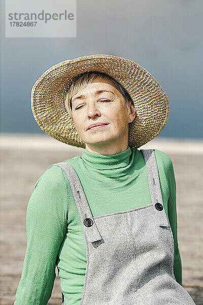 Reife Frau mit Hut sitzt an einem sonnigen Tag mit geschlossenen Augen