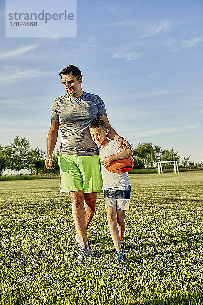 Lächelnder Vater und Sohn gehen an einem sonnigen Tag gemeinsam auf dem Sportplatz spazieren