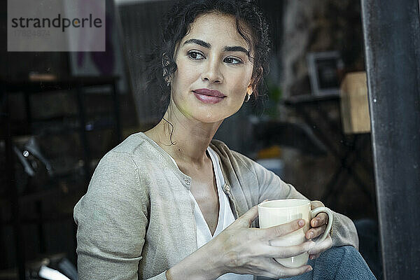 Lächelnde Frau mit Kaffeetasse  die durch das Fenster schaut