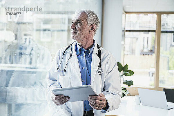 Nachdenklicher Arzt mit Tablet-PC  der durch das Fenster im Büro schaut
