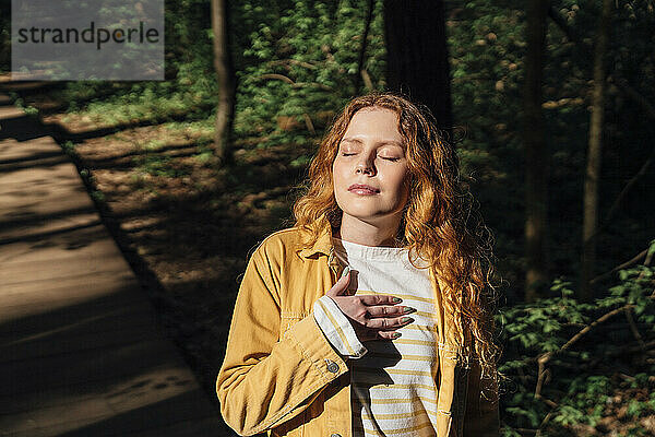 Young woman with hand on chest enjoying sunlight in forest