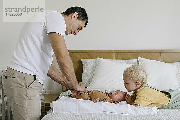 Junge blickt auf Vater  der auf dem Bett Babystrampler zuknöpft