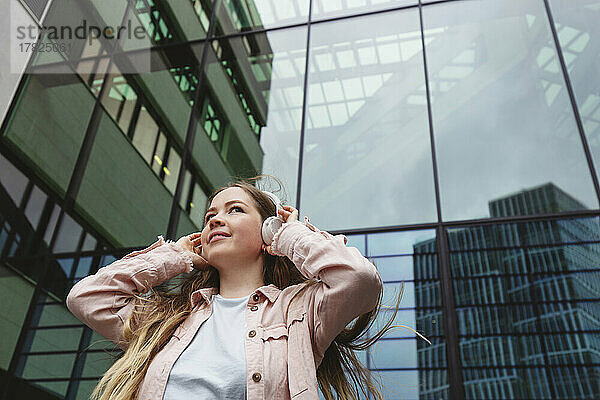 Lächelnde Frau mit kabellosen Kopfhörern steht vor dem Gebäude