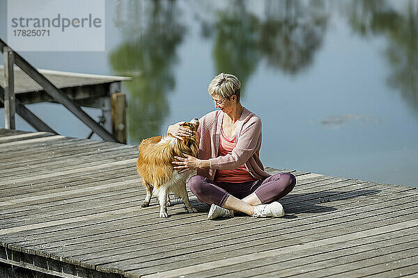 Reife Frau streichelt Hund  der auf dem Pier am See sitzt