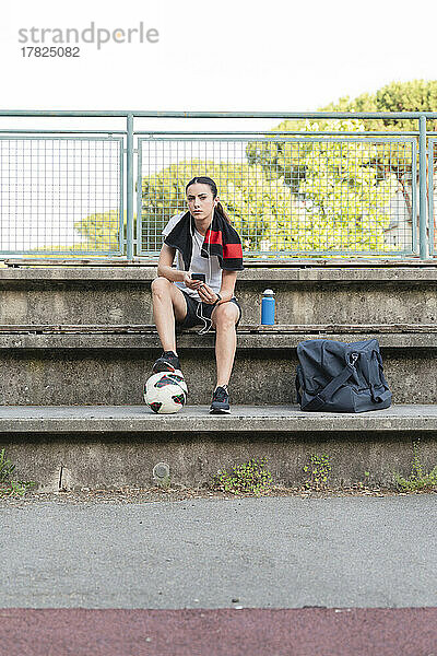 Frau hört Musik über In-Ear-Kopfhörer und sitzt mit Fußball auf Bank