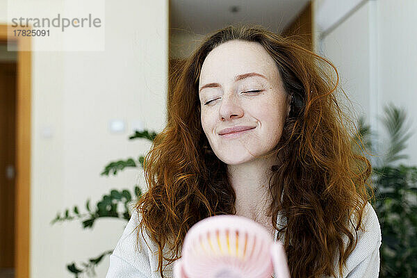 Lächelnde Frau mit geschlossenen Augen hält tragbaren Ventilator zu Hause