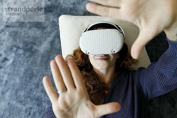 Glückliche Frau liegt mit VR-Headset auf dem Teppich und gestikuliert zu Hause