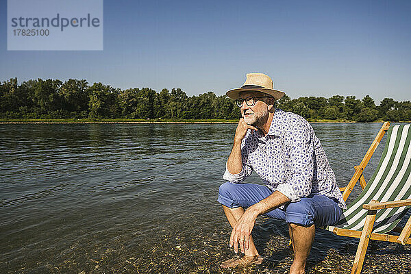 Mann mit Hut sitzt auf Liegestuhl am Flussufer