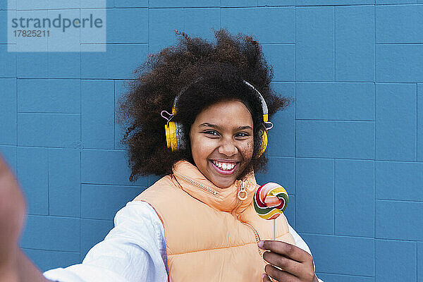 Fröhliches Mädchen mit Süßigkeiten  das über kabellose Kopfhörer vor der blauen Wand Musik hört