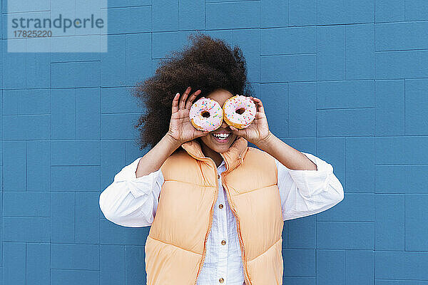 Glückliches Mädchen  das die Augen mit Donuts bedeckt und vor einer blauen Wand steht