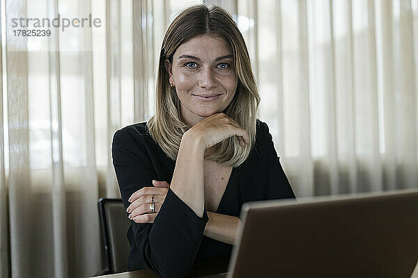 Lächelnde Geschäftsfrau sitzt mit der Hand am Kinn am Schreibtisch