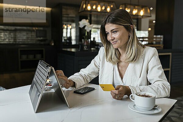 Lächelnde Geschäftsfrau mit Kreditkarte beim Online-Einkauf per Laptop im Restaurant