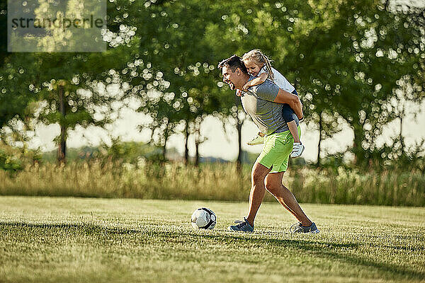Fröhliches Mädchen genießt die Huckepackfahrt eines Mannes  der auf dem Sportplatz Fußball spielt