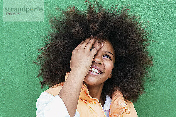 Glückliches Mädchen mit Afro-Frisur  das das Auge vor der grünen Wand bedeckt
