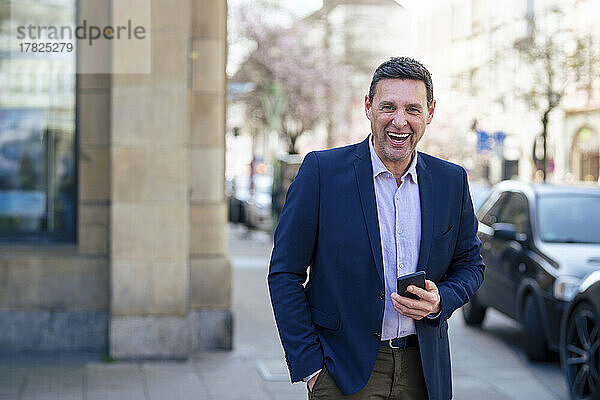 Geschäftsmann hält Smartphone lachend auf Fußweg