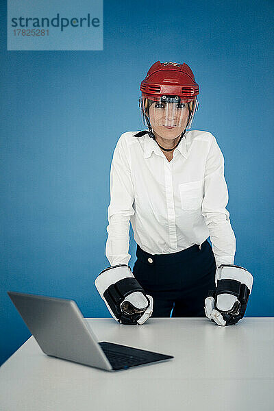 Geschäftsfrau mit Eishockeyhelm und Handschuhen am Tisch vor blauer Wand