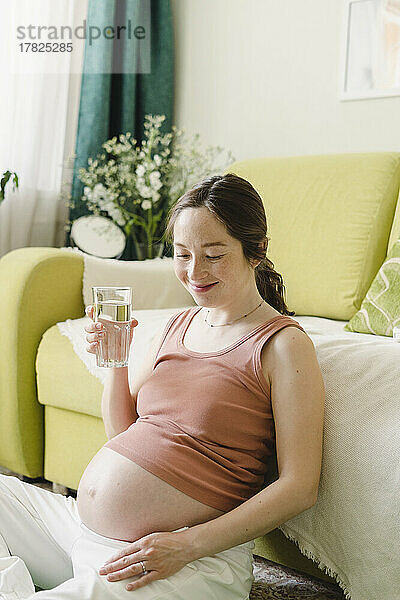 Lächelnde Frau mit einem Glas Wasser sitzt zu Hause auf dem Sofa