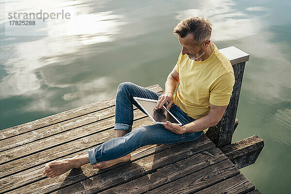 Reifer Mann sitzt mit Tablet-PC am Pier über dem See