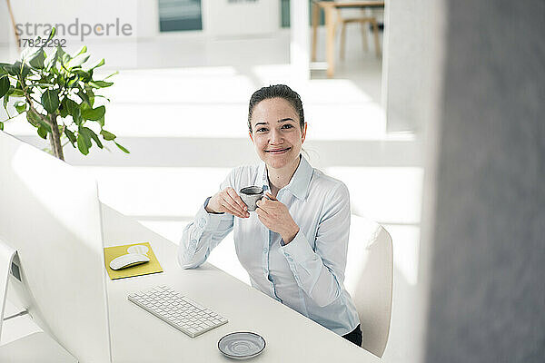 Lächelnde Geschäftsfrau mit Kaffeetasse am Schreibtisch im Büro