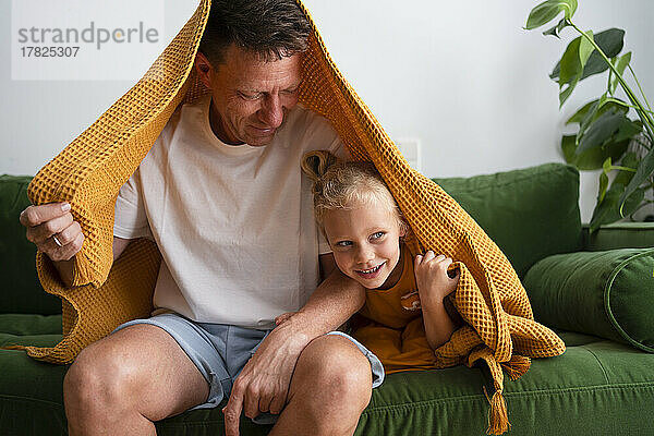 Vater und Tochter sitzen zu Hause unter einer Decke auf dem Sofa