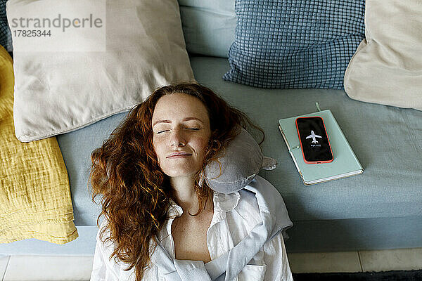 Lächelnde rothaarige Frau stützt sich mit dem Kopf auf das Smartphone und entspannt sich zu Hause auf dem Sofa